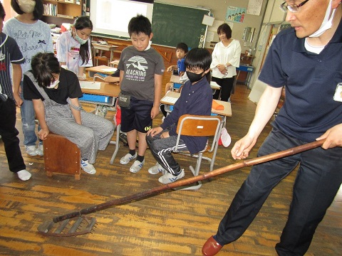 先生や児童たちが見ているなか、男性職員が細い竹で作られた先に櫛の様な形をし歯が4列付いている除草機の道具を使って除草の手本を見せている様子の写真