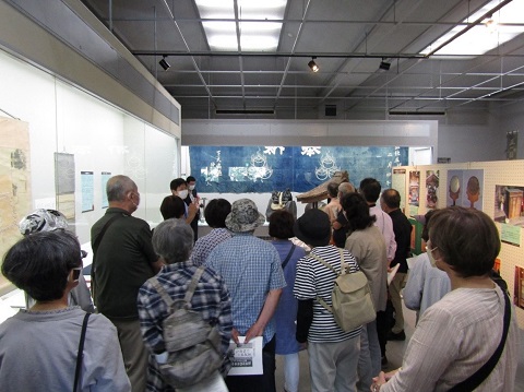 壁に様々な作品が展示され、前方に立つ福澤学芸員の説明を参加者が熱心に聞いている後ろ姿の写真