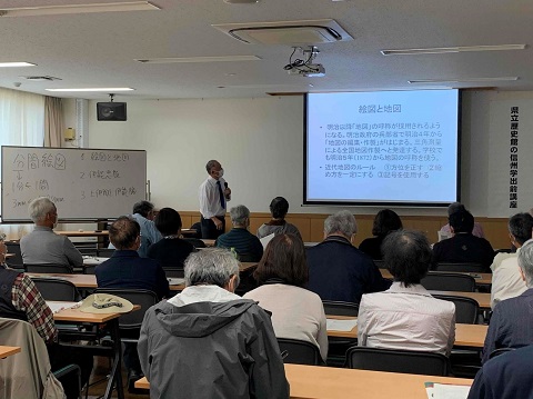 資料が映し出された前方のスクリーンの左側に立っている小野氏の話を、席に座った参加者が聞いている後ろ姿の写真