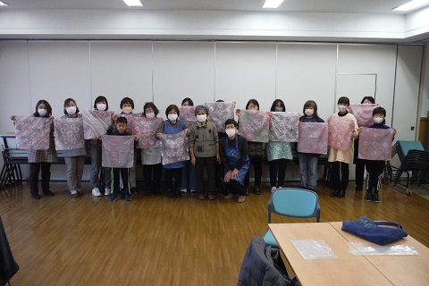 講師の林さんと女性スタッフを中央に、完成したハンカチを広げた草木染めに参加した15名の参加者が並んでいる記念写真