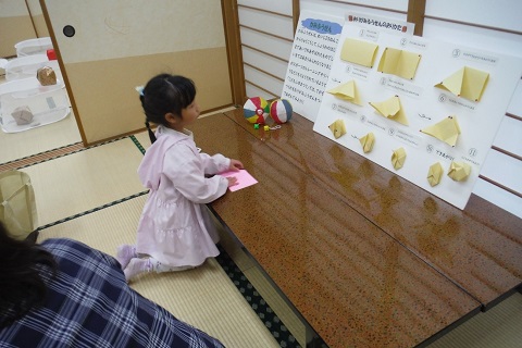 長机の上に置かれた紙ふうせんの折り方を見ながら折り紙を折っている、ピンク色の服を着た女の子の写真