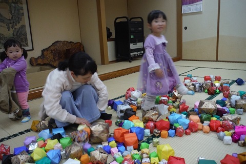 畳敷きの床に置かれた沢山の紙ふうせんの間を器用に歩いている女の子と、上着を脱ぎながら見ている小さな女の子と母親の写真