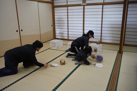 畳敷きの床に置かれた3つのカゴに向かって、2名の子供と母親が紙ふうせんをうちわで扇いで入れようと競争している様子の写真