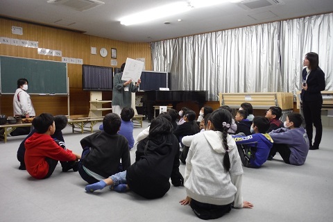 屋内の教室で前方に大きなボードを持った職員が立ち、床に座った児童たちが見ている出前授業の様子の写真