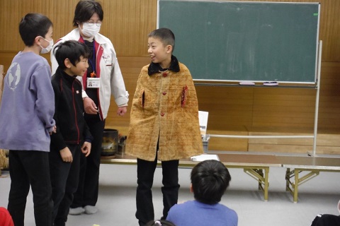 白色のジャンパーを着た女性職員と他の児童が見ているなか、子供用のマントを着た男児が一緒に立っている2名の男児を笑顔で見ている写真