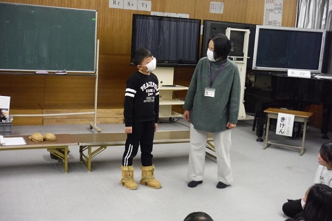 緑色の服を着た女性職員と藁製の雪靴を履いた男子児童が向かいあって立ち、女性職員に感想を伝えている様子の写真