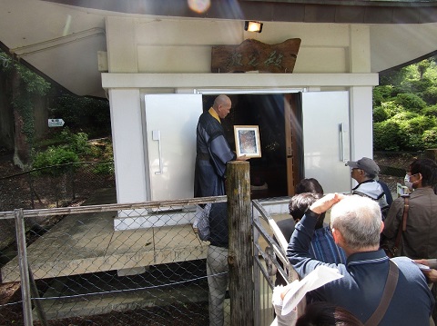 扁額に「放光殿」と書かれた建物の前で、額入りの写真を持つ住職の話を参加者が熱心に聞いている様子の写真