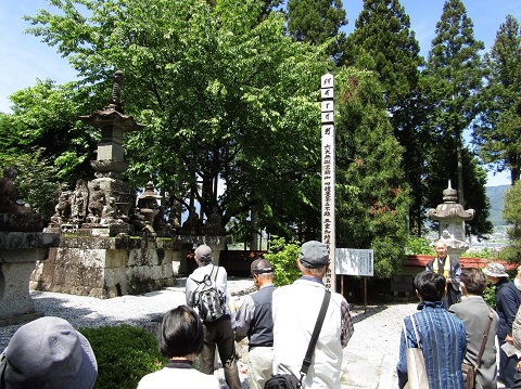 周囲を四天王、地蔵菩薩、不動明王などで囲んだ「宝篋印塔」の前に立つ住職の話を、参加者が熱心に聞いている後ろ姿の写真