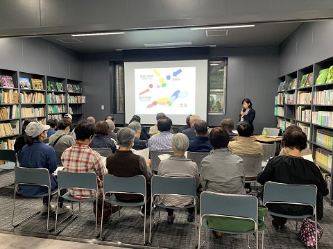 両側に本棚があり、前方にスクリーンが設置されている室内に参加者達が集まり、講師の水澤さんが室内前方で話をしている講座の様子の写真
