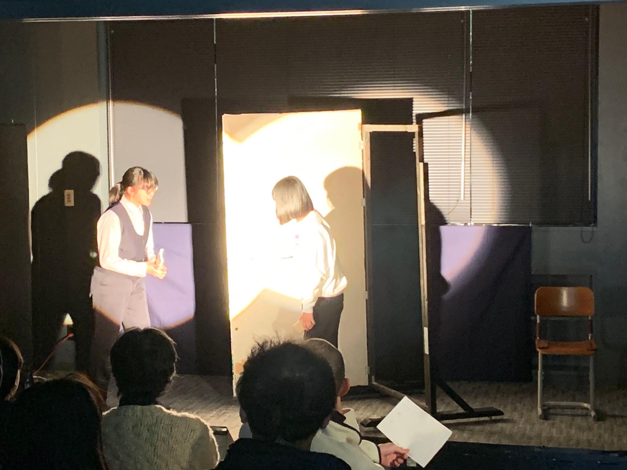 大道具のドアの前でスポットライトを浴びながら演技する2人の女子