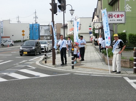 ランドセルを背負った小学生や、自転車に乗った学生が通学する横断歩道の前で、のぼり旗を持ち肩にタスキを掛けた3名の男性があいさつ運動を行っている様子の写真