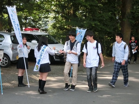 周囲に木々が生えている細い道路沿いで、制服姿の女子高生がのぼり旗を持って啓発活動を行っているなか、「信州あいさつ運動」と書かれたタスキを肩に掛けた女子学生が道を歩いている若い男性に啓発物品などを配布している街頭啓発活動の様子の写真
