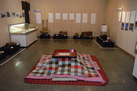 壁に資料や写真、着物が展示され、ショーケースや生活道具などが並んだ中央に、チェック柄の布団が敷かれたこたつが設置されている写真