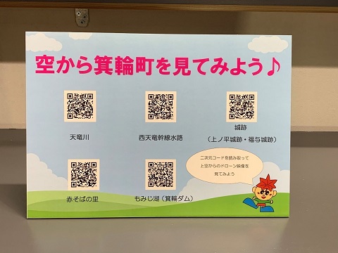 「空から箕輪町を見てみよう」と書かれたボードに、町内の主要地の5か所をドローンで撮影した空の映像が見られる二次元コードが表示されている写真