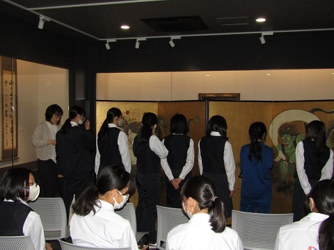 左側に女性職員が立ち、展示されている「風神雷神図屏風・夏秋草図屏風」の前に7名の女子高生が並んで立ち、鑑賞している様子の写真