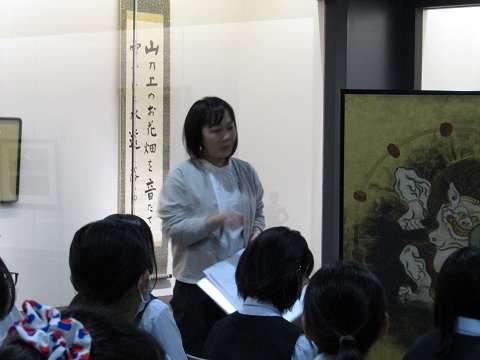 展示されている「風神雷神図屏風・夏秋草図屏風」の左側に立つ中村さんの話を、椅子に座って聞いている女子高生の後ろ姿の写真