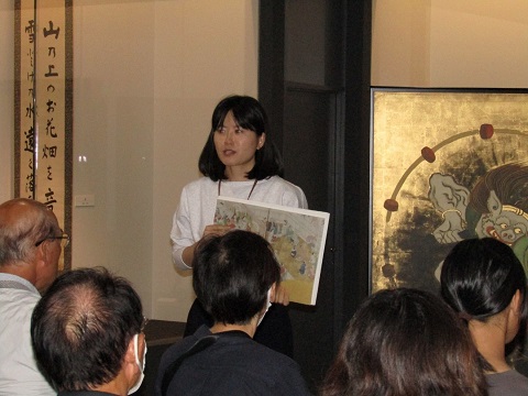 展示されている「風神雷神図屏風・夏秋草図屏風」の左側に立つ松沼さんが資料を持ちながら解説している様子を、椅子に座った参加者が聞いている後ろ姿の写真