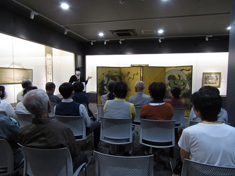 展示されている「風神雷神図屏風・夏秋草図屏風」の左側に立つ小島さんが手に資料を持ち解説している様子を、参加者が椅子に座り熱心に聞いている後ろ姿の写真