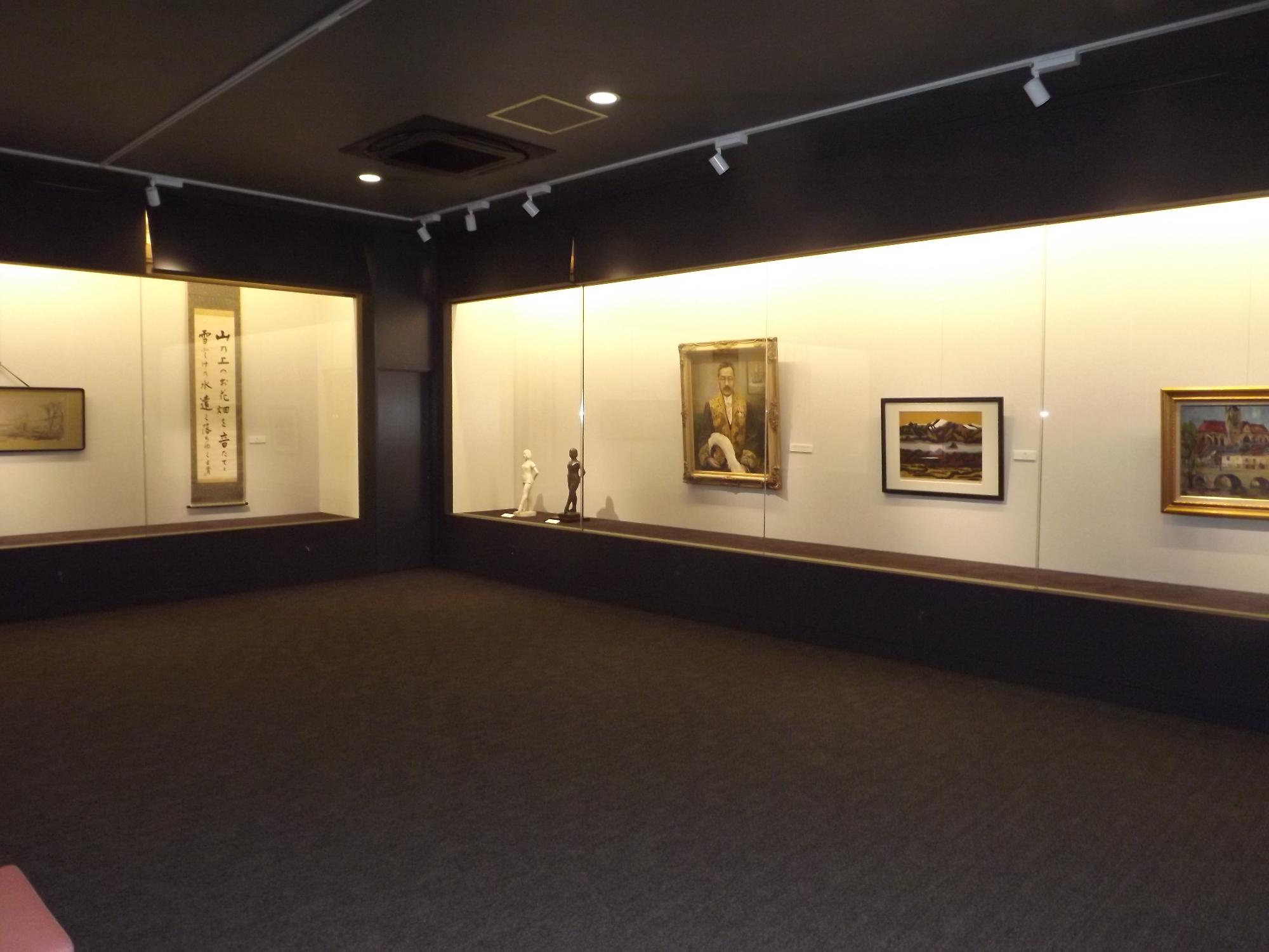 全体的に暗い美術展示室。展示ケースだけが明るく光る。中には書の掛け軸、女性の彫刻、男性の肖像画、山々の版画が展示されている
