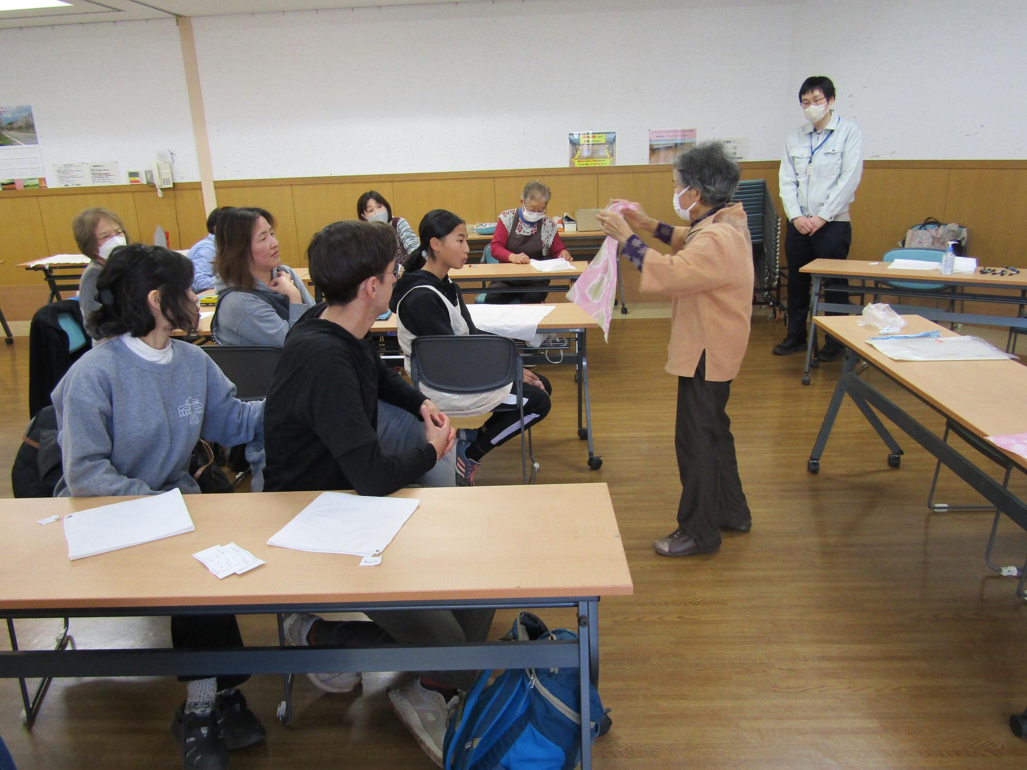 座っている受講生の前で草木染めをしたピンク色のハンカチを持って立ちながら説明する講師の林先生