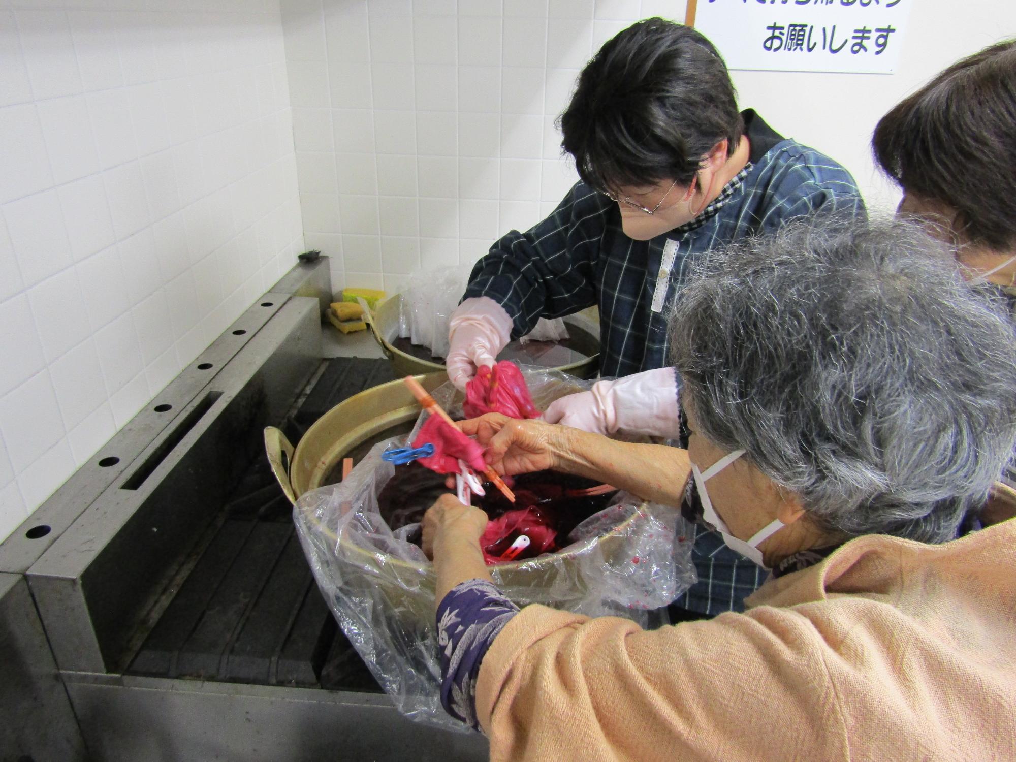 割り箸や洗濯ばさみをつけたままのハンカチを抽出剤につける講師たち。ハンカチは赤く染まっている
