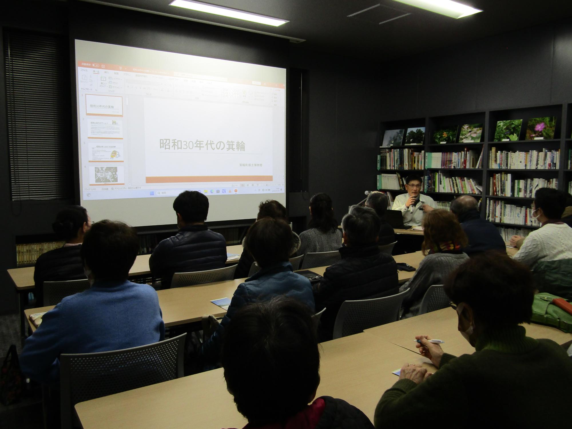 椅子に座った10名ほどの参加者が前のスクリーンを見ている。前では職員が説明している。