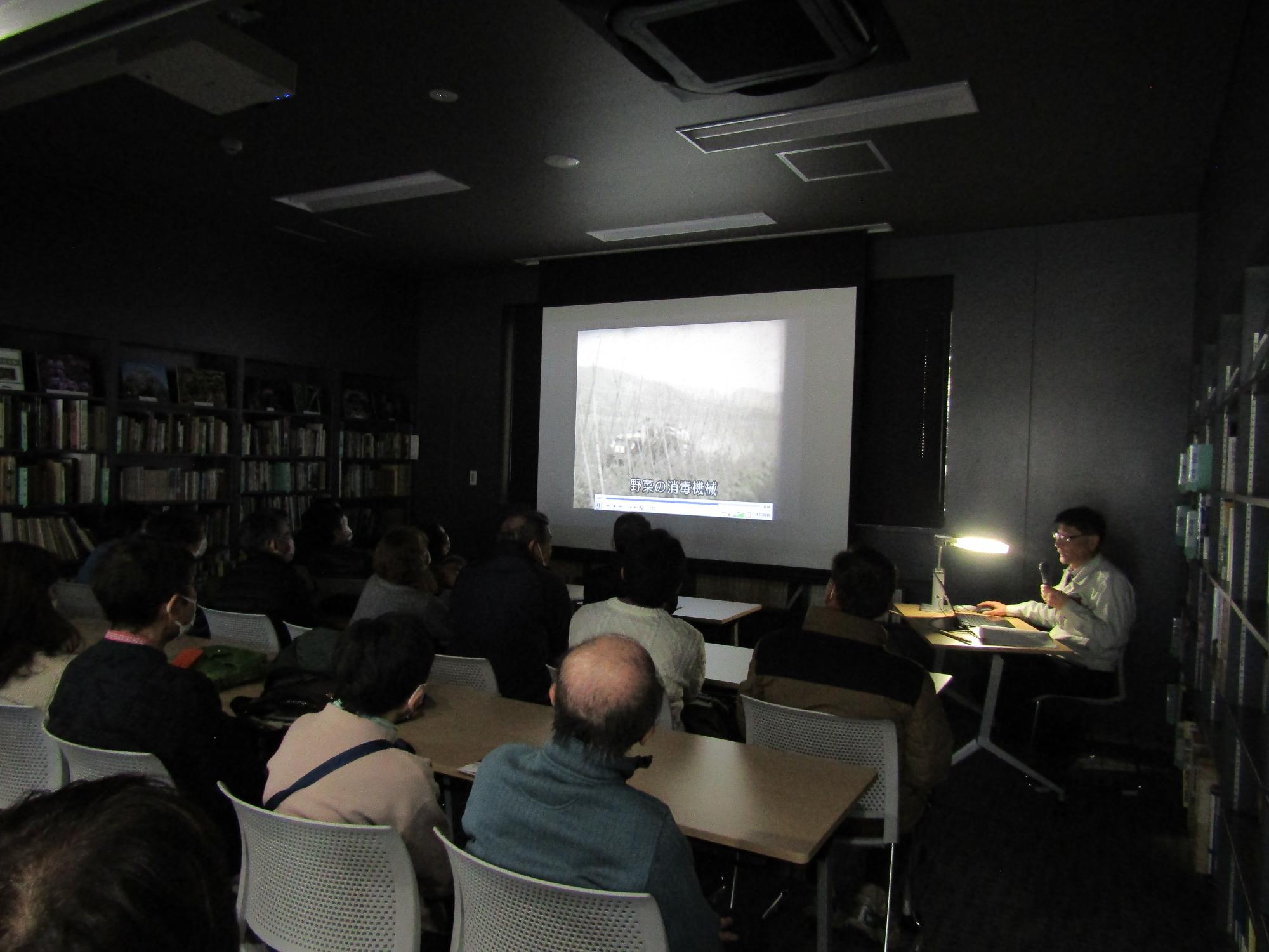 昔の映像を眺める参加者。室内は暗く、スクリーンに映像が映し出されている。