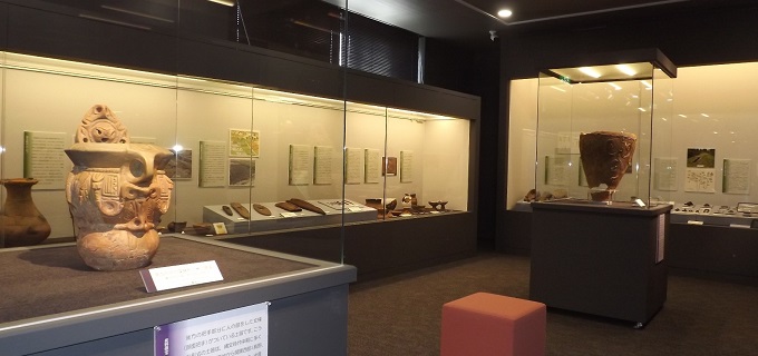 壁が黒色の歴史展示室。独立ケースには縄文土器が展示され、奥の展示ケースには弥生時代の土器や木製品、古墳時代の土器や古墳の副葬品、埴輪などが展示されている