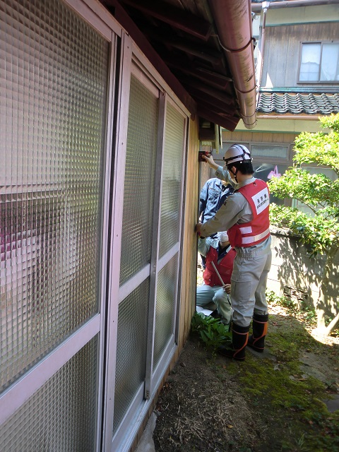 ヘルメットを被り赤いベストを着た作業員の方が住宅の屋外の壁にテープメジャーをあてて調査をしている様子の写真