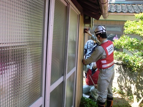 ヘルメットを被り赤いベストを着た作業員の方が住宅の屋外の壁にテープメジャーをあてて調査をしている様子の写真