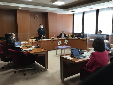 机がロの字に設置された室内に議員の方々が座り、前方の男性が立ち話をしている様子を聞いている視察の様子の写真