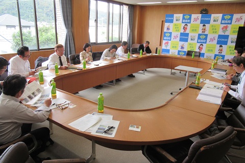 横長のO型の机の左側に議員の方々が座り、右側に座っている男性担当者の話を聞いている視察の様子の写真