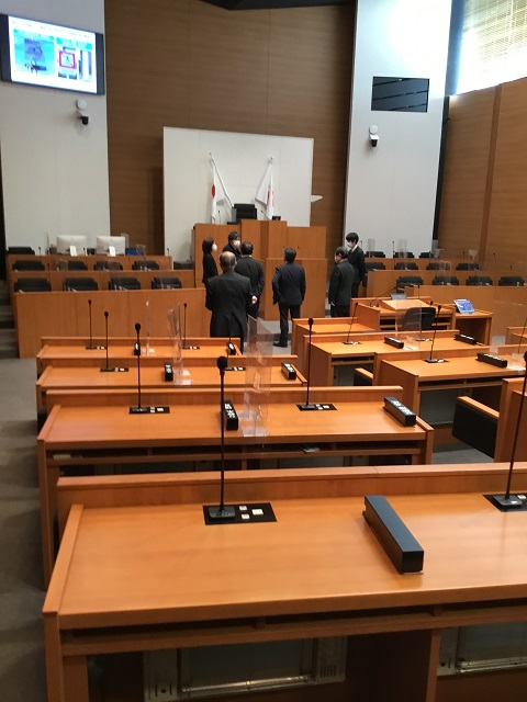天井が高く左壁上に大きなモニターが設置された議会室内で、7名の議員の方々が立っている視察の様子の写真
