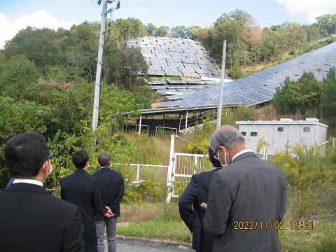 小高い山の上に設置された太陽光パネルを視察している議員の方々の写真