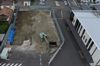フェンスで囲まれた中に緑色の重機が入って工事が進められている建設予定地を駐車場側の上空から撮影した写真
