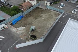 フェンスで囲まれた中に緑色の重機が入って工事が進められている建設予定地を敷地内に建つ建物側の上空から撮影した写真