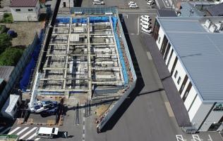 フェンスで囲まれた中に長く伸びたクレーン車が入り基礎工事が進められている建設予定地を駐車場側の上空から撮影した写真