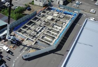 フェンスで囲まれた中に長く伸びたクレーン車が入り基礎工事が進められている建設予定地を敷地内に建つ建物側の上空から撮影した写真