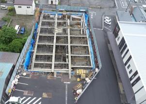 資材が置かれたフェンスの内側がブルーシートで囲まれ、基礎工事が進められている建設予定地を駐車場側の上空から撮影した写真