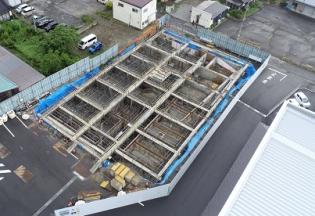 資材が置かれたフェンスの内側がブルーシートで囲まれ、基礎工事が進められている建設予定地を敷地内に建つ建物側の上空から撮影した写真