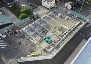 フェンスで囲まれた工事が進められている中に入り緑色の重機が工事を行っている建設予定地の様子を敷地内に建つ建物側の上空から撮影した写真
