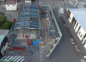 屋根部分の骨組みが取り付けられ赤色の重機で工事が進められている建設予定地の様子を駐車場側の上空から撮影した写真