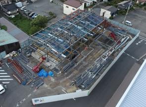 屋根部分の骨組みが取り付けられ赤色の重機で工事が進められている建設予定地を敷地内に建つ建物側の上空から撮影した写真