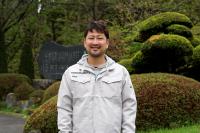 綺麗に手入れされた植木の前に立つ、作業着を着た笑顔の柴田さんの上半身の写真