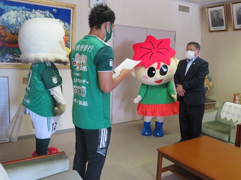 ガンズくんと緑色のユニフォームを着た男性が、もみじちゃんとスーツを着た男性と室内で対面している写真