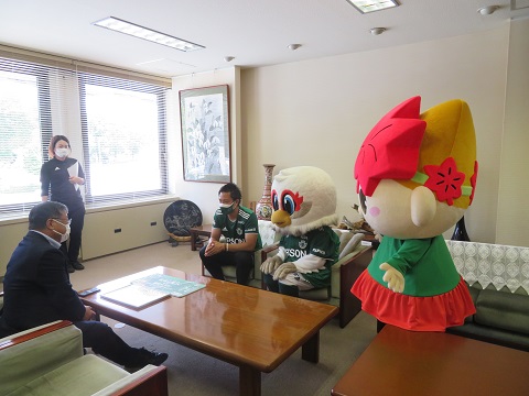 緑色のユニフォームを着た男性とガンズくんがスーツを着た男性と座って対面している写真