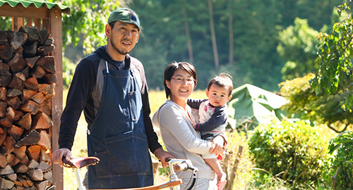 屋外で自転車と一緒に写っている安田さんと男の子を抱いている奥様の写真（移住者ファイル2安田さんちのページへリンク）