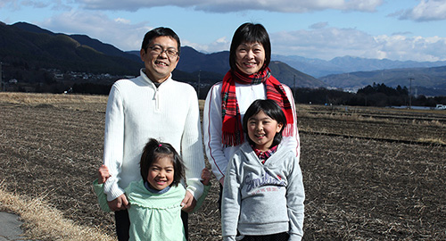 遠くに見える山々と広々とした畑の前に立っている城内さんと奥様、二人の子供の写真（移住者ファイル5城内さんちのページへリンク）