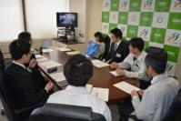 長机の席について前方のモニター画面を見ながら話し合いをしている会議の参加者の写真