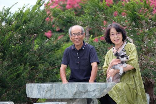 屋外の石でできたテーブルとイスに腰掛けている等々力さんと犬を抱いた奥様の写真